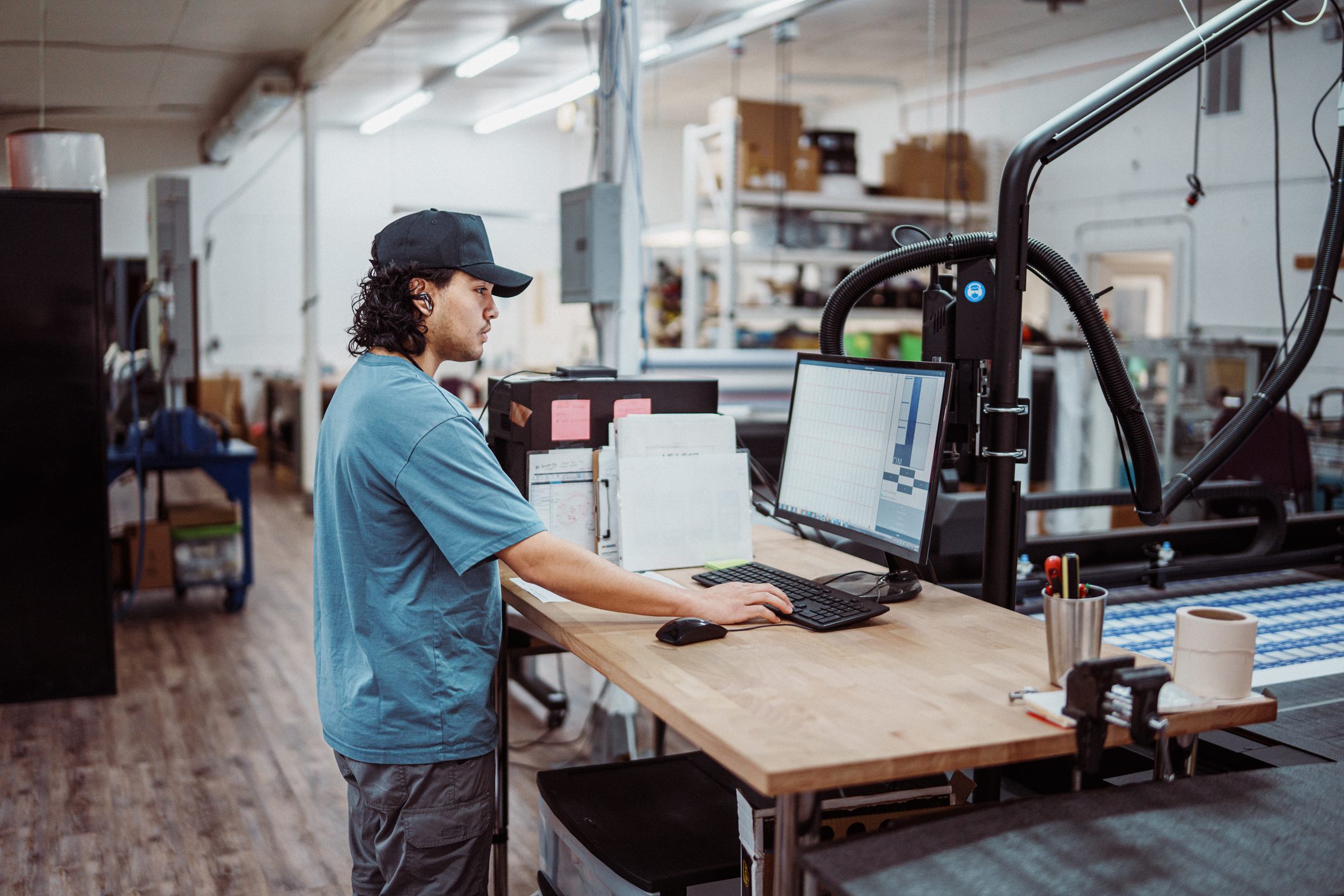 A male worker uses a computer when adjusting the settings on a large format printer and paper cutter in a manufacturing and fabrication facility.